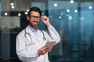 Portrait of a young handsome male doctor. He stands in the office in a white coat and with a stethoscope. He holds a tablet in his hands. He looks at the camera, adjusts his glasses, smiles. - Powered by Adobe