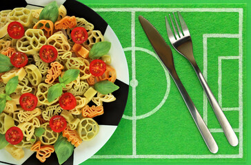 Fototapeta na wymiar Tricolor pasta in form of football boots, balls, cups, green basil, red cherry tomato on plate like soccer ball, dish mat like football field and cutlery: knife and fork. Italian cuisine concept.