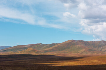 Motley autumn landscape with sunlit high mountain plateau and mountain range under dramatic cloudy sky. Vivid autumn colors in mountains. Sunlight and beautiful shadows of clouds in changeable weather