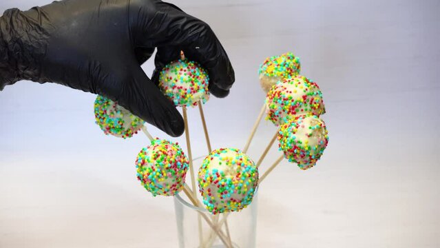 Bright cake-pops candy on stick on white background. Proffessional baker in black gloves serves sweets, party candy bar. Kids bisquit balls dessert covered by chocolate, colored confectionery topping
