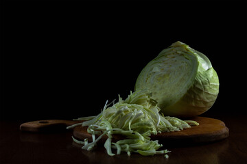 white cabbage is cut and finely chopped on a wooden cutting board on a table against a dark background. side view. moody artistic still life in simple rustic style with copy space