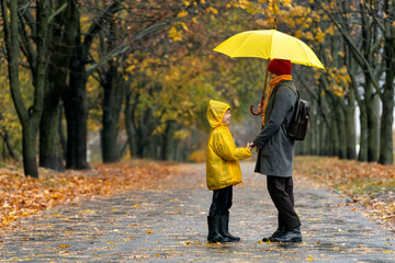 Son with mom walking in the autumn park in rain with large yellow umbrella. Rainy family day