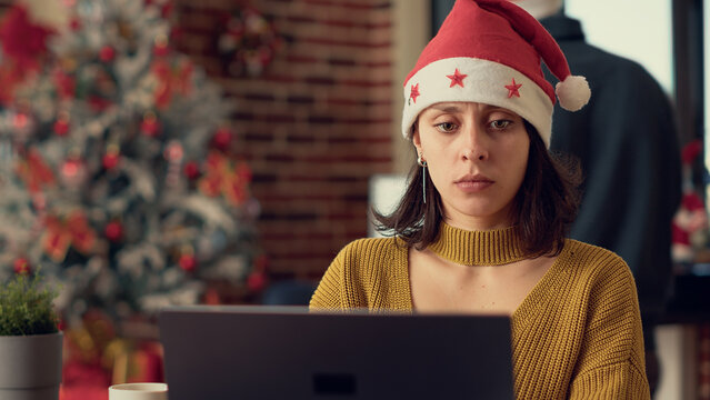 Female Employee Analyzing Research Data On Laptop, Wearing Santa Hat During Christmas Eve Holiday Season. Working On Computer To Plan Report Before Celebrating Winter Event. Tripod Shot.