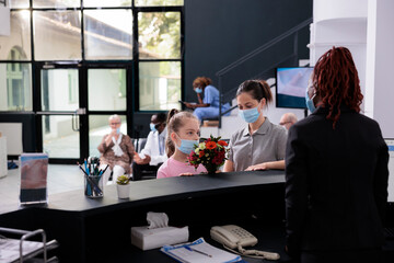 Mother standing at reception counter with child discussing with receptionist during checkup visit appointment in hospital lobby. People wearing medical face mask to prevent infection with covid19