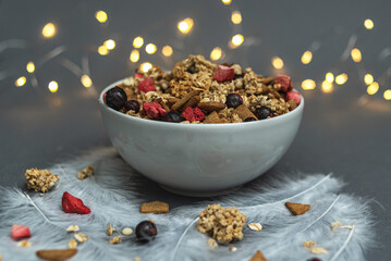 A bowl of muesli on a gray monochrome background, with white feathers and a side from a garland. Healthy food, healthy breakfast