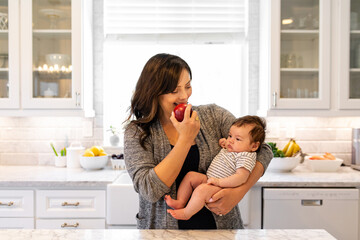 Woman Eats A Healthy Snack While Holding Newborn Baby In The Kitchen