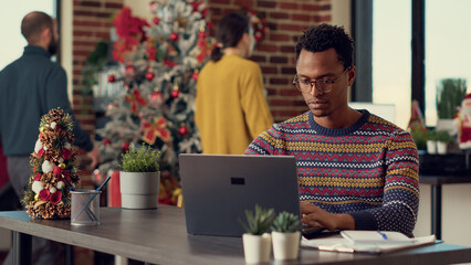 Company employee using laptop in christmas decorated office filled with tree and lights for winter festivity. Young adult working with documents at desk during xmas holiday season.