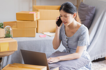 Smiling beautiful Asian woman SME entrepreneur Business owner holding cardboard boxes pack products and check customers' orders on their laptops at home, concept of SME delivery online.