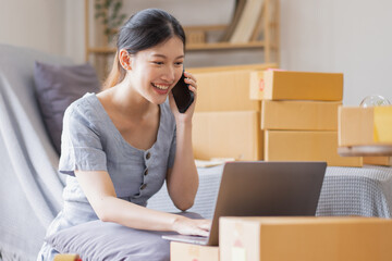 Fototapeta na wymiar Smiling beautiful Asian woman SME entrepreneur Business owner holding cardboard boxes pack products and check customers' orders on their laptops at home, concept of SME delivery online.