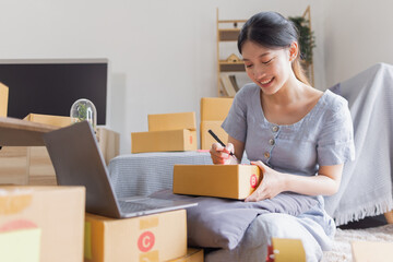 Smiling beautiful Asian woman SME entrepreneur Business owner holding cardboard boxes pack products and check customers' orders on their laptops at home, concept of SME delivery online.