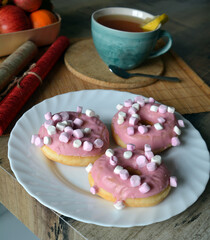 Cup of black tea with lemon and three donuts with pink icing on a plate on a wooden table. Sweet breakfast.