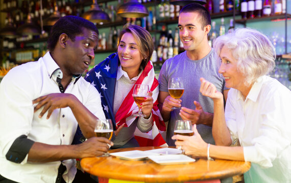 Young and senior football and soccer fans drinking beer at pub, cheering and celebrating scores. Girl with american flag on her shoulders