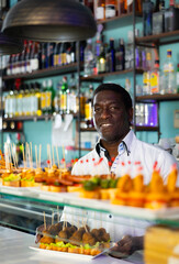 Smiling African american man bartender offering tasty pinchos to customer in cafe