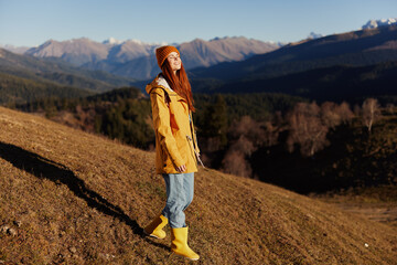 Naklejka premium Woman smile with teeth full-length walk happiness walking on the hill and looking at mountains in a yellow raincoat and jeans in the autumn happy journey in the sun sunset hiking, freedom lifestyle 