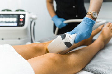 Obraz premium SPA and Beauty Salons treatment - laser hair removal from legs. Unrecognizable caucasian beautician in protective blue gloves using laser on her caucasian client. Hair removal cosmetology procedure