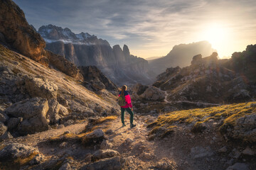 Fototapeta premium Girl with backpack on the trail in mountains at sunset in autumn. Beautiful landscape with young woman, high rocks, path, stones, orange grass, sky with clouds in fall in Dolomites, Italy. Adventure