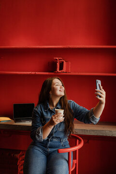 Young cheerful freelancer woman taking selfie in cafe with coffee cup