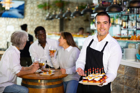 Smiling waiter posing and holding tray with order at beer bar with customers his behind