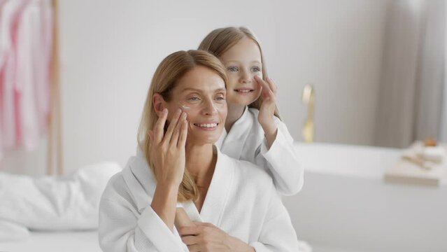 Beauty day at home. Positive mother and her little daughter applying pampering cream on faces, free space