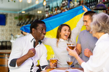 Diverse group celebrating with Ukrainian flag at a pub with beer