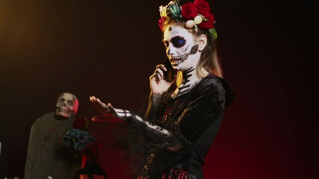 Spooky goddess talking on phone call with smartphone, wearing flowers crown and cavalera catrina halloween costume. Woman dressed as santa muerte to celebrate mexican culture holiday. Handheld shot.