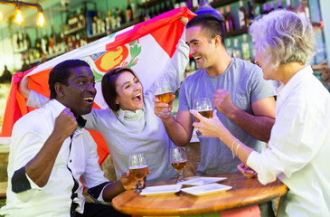 Excited diverse soccer fans with flag of Peru rejoicing winning game with glasses of beer in the pub