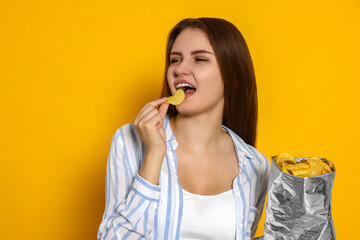 Pretty young woman eating tasty potato chips on yellow background
