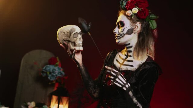 Santa muerte model acting flirty with skull and black roses, wearing traditional body art to celebrate mexican holiday. Looking like goddess of death on holy dios de los muertos ritual. Handheld shot.