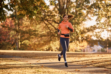 Full length of happy sportswoman jogging in autumn park. - obrazy, fototapety, plakaty