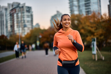 Happy sportswoman with earbuds running in park. - obrazy, fototapety, plakaty