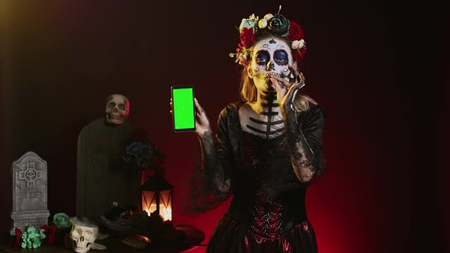 Creepy female model holding greenscreen on smartphone, using mobile phone with isolated mockup template. Woman dressed as santa muerte showing blank chroma key copyspace display.