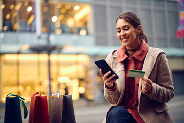 Happy woman using smart phone and credit card while shopping in city.