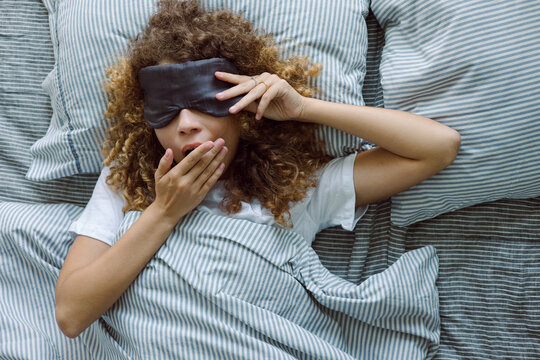 Portrait of curly woman in sleep mask yawning in bed