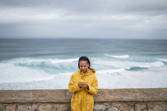 young woman with mobile phone and yellow raincoat by the sea