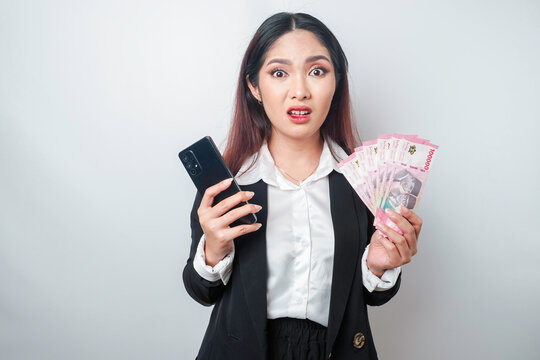 A confused young businesswoman is wearing a black suit, holding her phone and money in Indonesian rupiah isolated by a white background