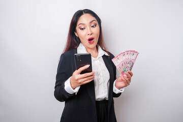 Surprised Asian businesswoman wearing black suit holding her smartphone and money in Indonesian Rupiah, isolated by white background