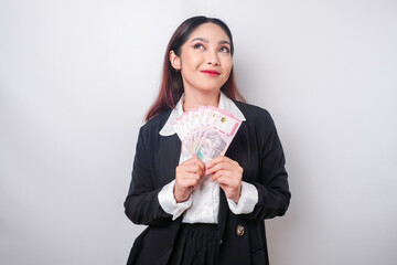 A thoughtful young woman is wearing black suit and holding cash money in Indonesian rupiah isolated...