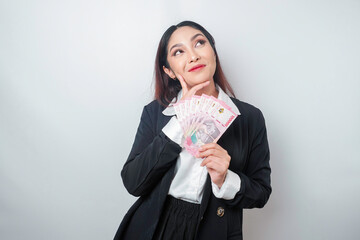 A thoughtful young woman is wearing black suit and holding cash money in Indonesian rupiah isolated...