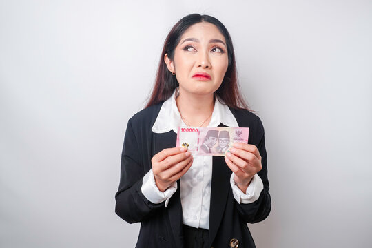 A thoughtful young woman is wearing black suit and holding cash money in Indonesian rupiah isolated by white background
