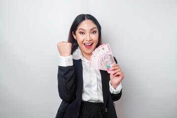 A young Asian businesswoman with a happy successful expression wearing black suit and holding money in Indonesian Rupiah isolated by white background