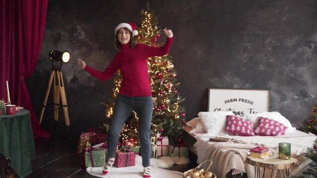 Woman in red sweater and Santa's hat dancing near the Christmas tree