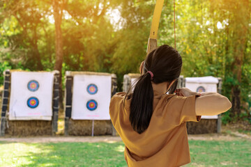 Back view of Asian girl wear face mask aims archery bow and arrow to colorful target in shooting...