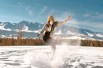 Happy girl in sweater and knitted hat is having fun and kicks snow in mountains
