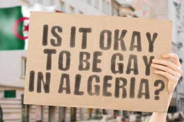 The question " Is it okay to be gay in Algeria? " is on a banner in men's hands with blurred background. Friendly. Passionate. Contact. Date. Dating. Lover. Partner. Boyfriend. Pleasant. Approval