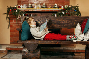 A charming baby is lying on a bench with a book of fairy tales in her hands against the background of a fireplace decorated for Christmas holidays. New Year, Christmas, family holiday