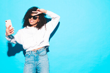 Beautiful black woman with afro curls hairstyle. Smiling model dressed in white summer top and jeans clothes. Sexy carefree female posing near blue wall in studio. Tanned and cheerful. Takes selfie