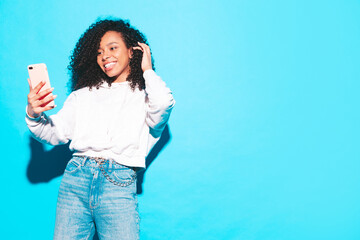 Fototapeta premium Beautiful black woman with afro curls hairstyle. Smiling model dressed in white summer top and jeans clothes. Sexy carefree female posing near blue wall in studio. Tanned and cheerful. Takes selfie
