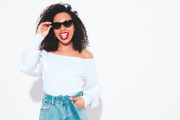 Naklejka premium Beautiful black woman with afro curls hairstyle. Smiling model dressed in summer top and skirt clothes. Sexy carefree female posing near blue wall in studio. Tanned and cheerful. Isolated on white