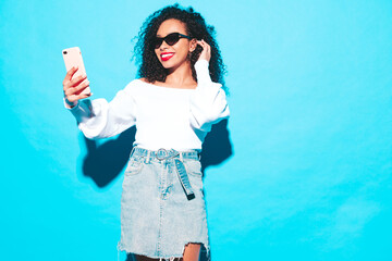 Beautiful black woman with afro curls hairstyle. Smiling model dressed in white summer top and jeans clothes. Sexy carefree female posing near blue wall in studio. Tanned and cheerful. Takes selfie