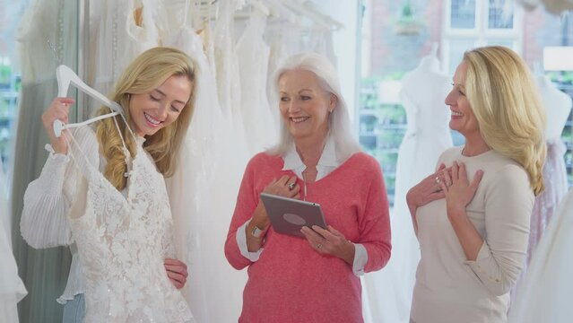 Sales assistant or shop owner with digital tablet helping mother with adult daughter in bridal store to choose wedding dress - shot in slow motion